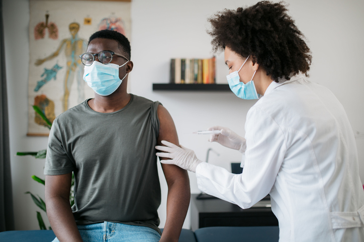 Young African American male patient sitting in a medical clinic and is being given the Covid 19 vaccine in his shoulder by a female African American doctor, both wearing protective face masks